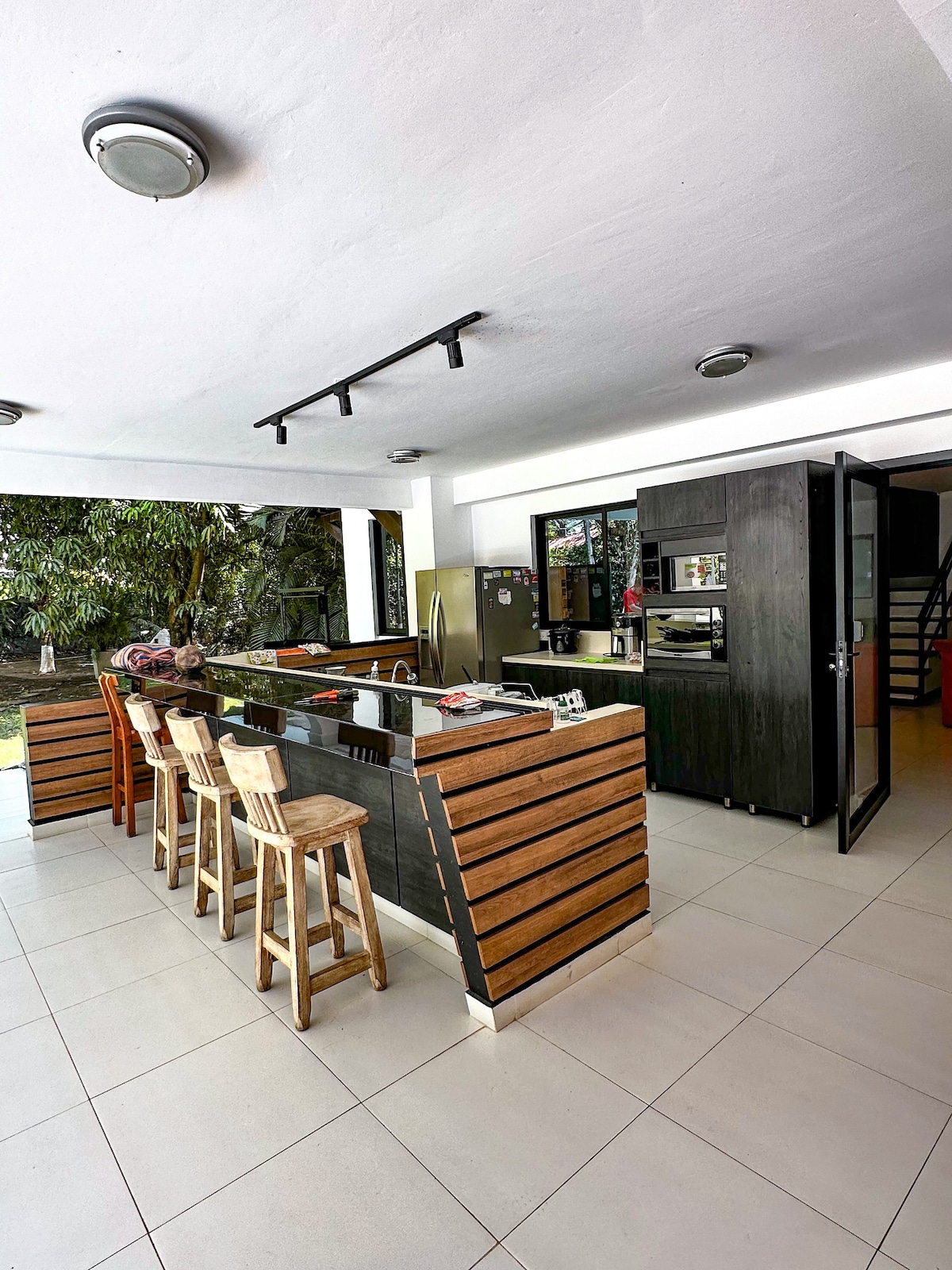 A spacious kitchen area features a central island with bar stools, positioned next to large glass doors leading to a lush outdoor space. Modern appliances, including a refrigerator and oven, are visible, providing a welcoming environment for meal preparation and gatherings.