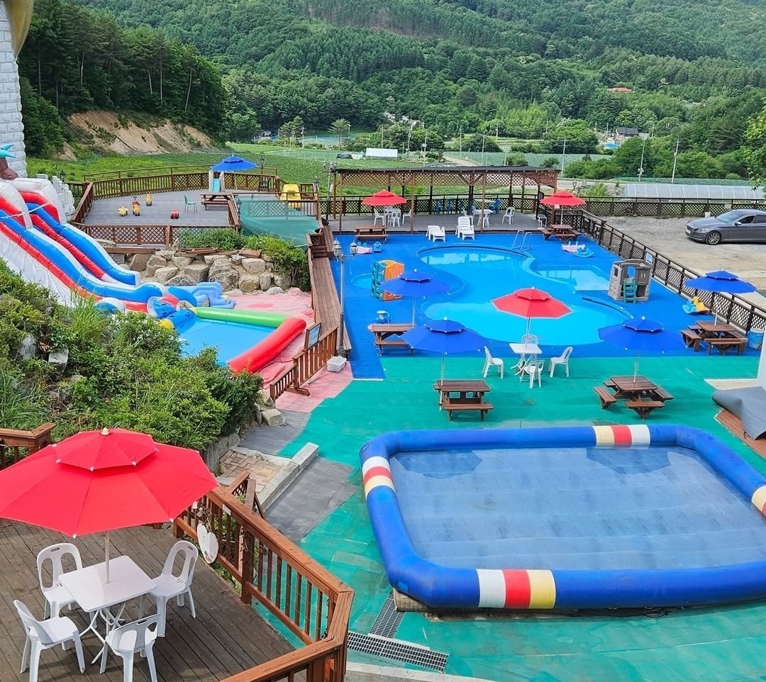 A vibrant outdoor area features a large swimming pool with colorful patterns, alongside several water slides. Brightly colored umbrellas provide shade over picnic tables. Lush greenery and hills are visible in the background, contributing to a refreshing atmosphere.