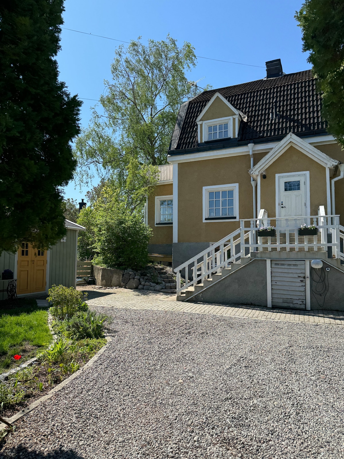 A charming three-story yellow house is shown, featuring a slate roof and white-framed windows. A welcoming front entrance with a staircase leads up from the gravel driveway, surrounded by garden areas with greenery and flower beds.