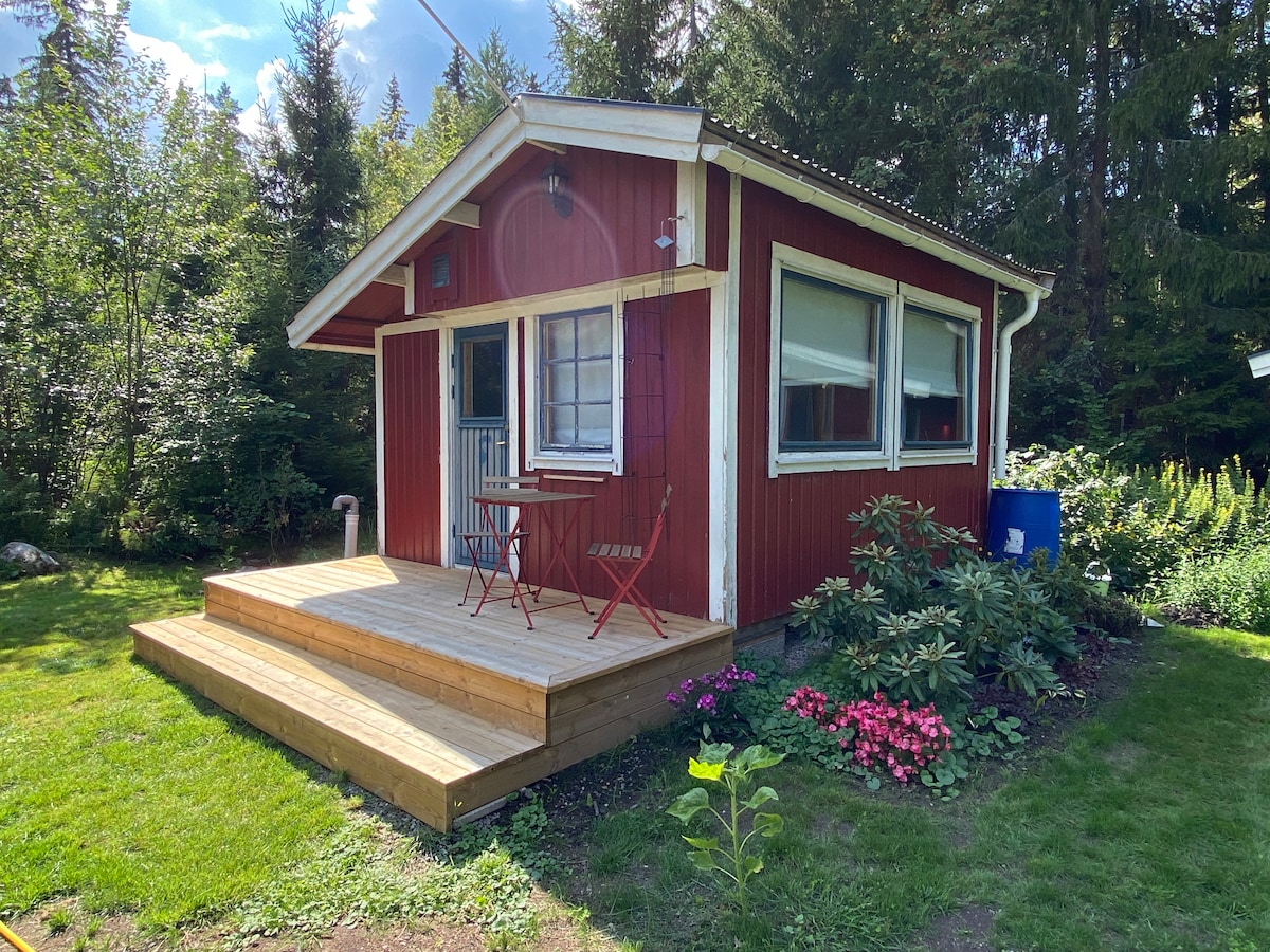 A red cottage is framed by lush greenery, featuring a wooden deck with two folding chairs and a small table. Flower beds with colorful blooms are visible, enhancing the tranquil outdoor setting. Large windows allow natural light into the inviting space.