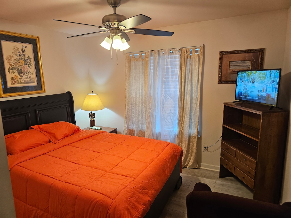 A bright bedroom features an orange quilted bedspread atop a spacious bed. A nightstand with a lamp complements the decor. Two light-filtering curtains frame the window, and a television is positioned on a wooden dresser, providing entertainment options.