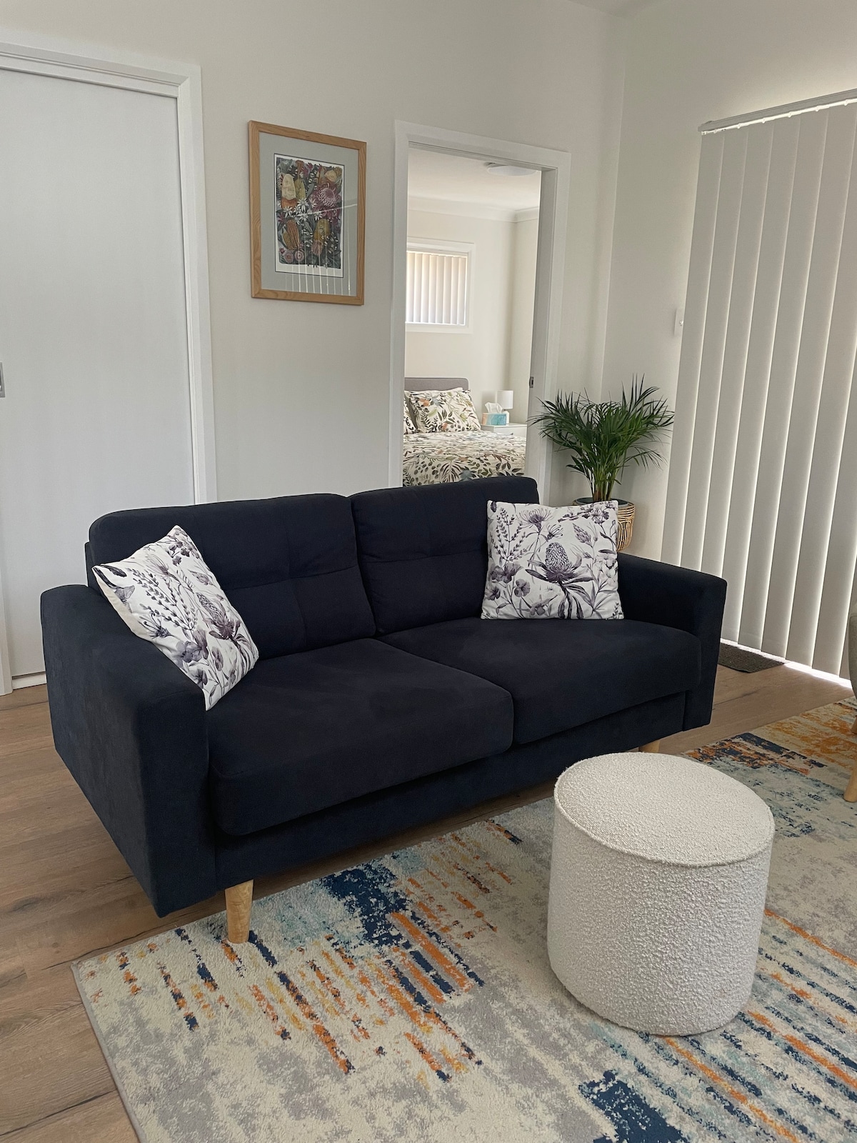 A modern living area features a dark sofa adorned with decorative cushions, positioned in front of a small round white table. A colorful rug adds texture to the floor, while natural light filters in through vertical blinds, brightening the space.