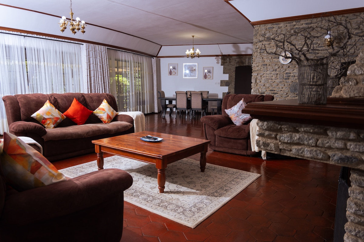 A spacious living room is featured, furnished with comfortable sofas adorned with colorful cushions. A wooden coffee table is centered on a patterned rug, while a stone fireplace adds texture to the room. Large windows allow natural light to brighten the space, and a dining area is visible in the background.