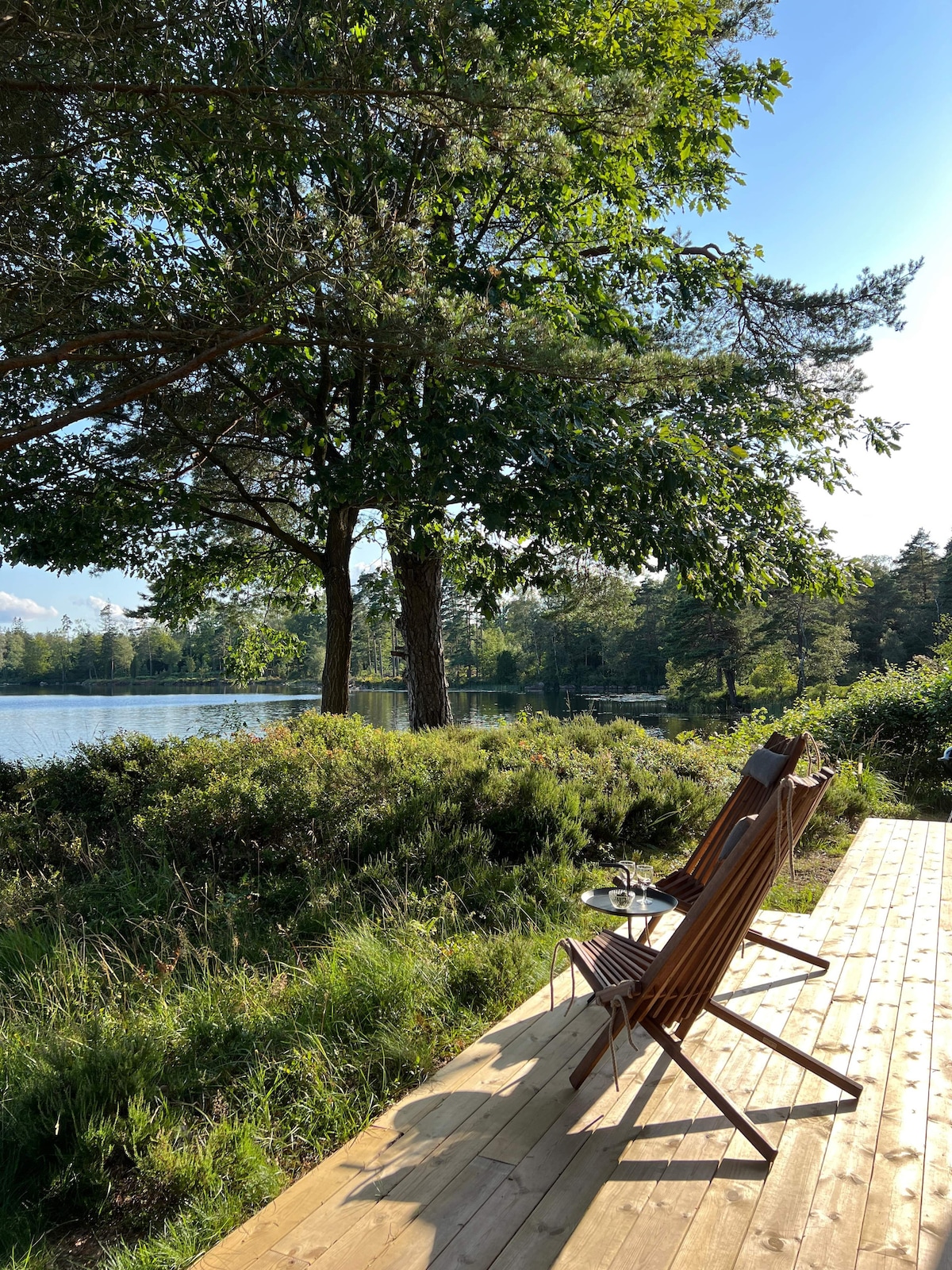 Two wooden lounge chairs are positioned on a spacious wooden deck overlooking a serene body of water. Lush green trees frame the scene, providing shade and a natural ambiance. The gentle ripples of the water reflect the clear sky and surrounding landscape.