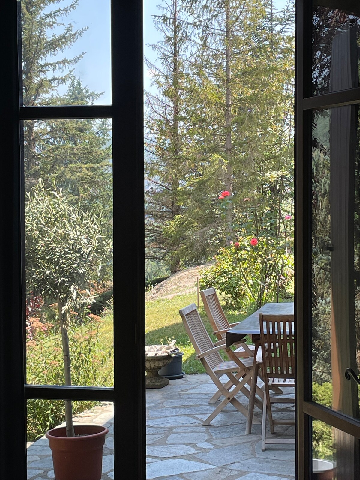 A serene outdoor patio is visible through a large window, showcasing wooden furniture arranged around a stone surface. Lush greenery surrounds the area, with flowering plants adding color to the scene. A terracotta pot with a small plant is placed near the window.
