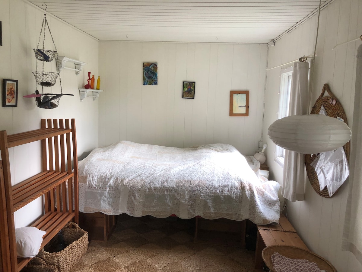A cozy sleeping area is defined by a spacious bed covered with a light quilt. Shelves with decorative items are mounted on the white walls, and simple artwork adorns the room. A warm light fixture complements natural light filtering through the sheer curtains.