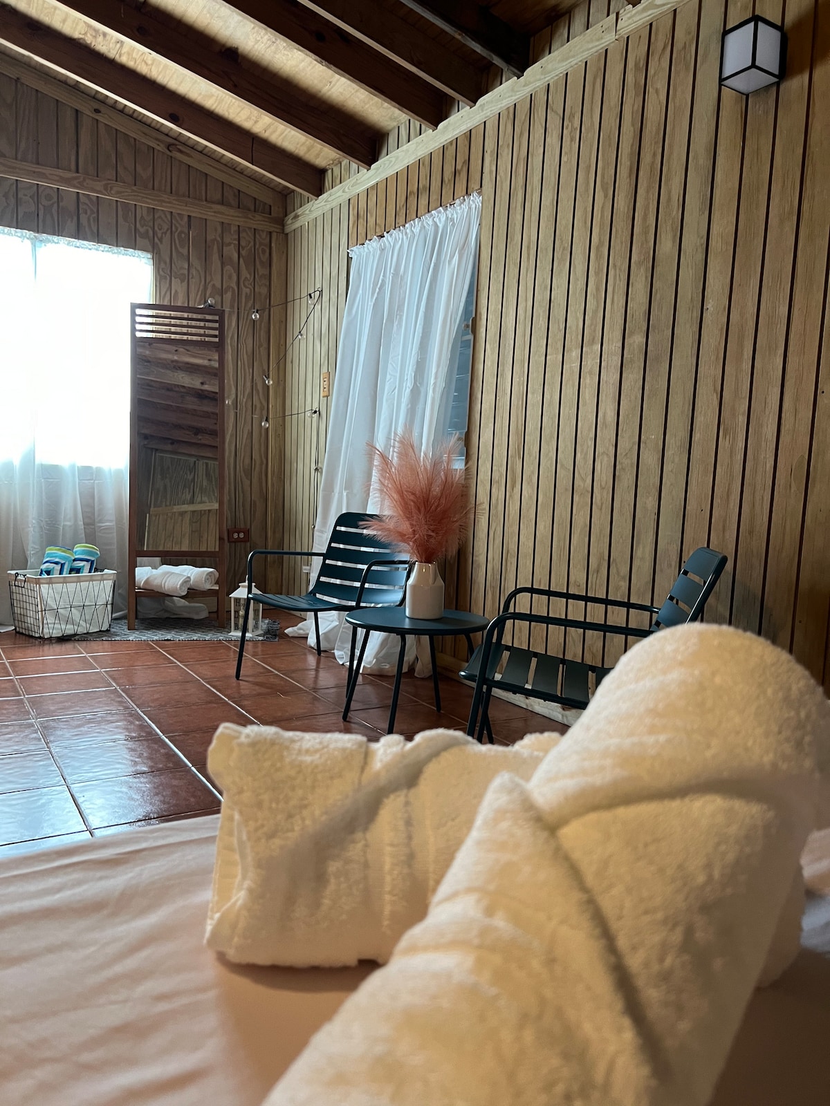 A cozy interior space features wooden walls and a tiled floor. Two chairs are positioned near a window, which is dressed with light curtains. Fresh towels are neatly arranged on the bed, with a vibrant plant in a vase accentuating the serene atmosphere.