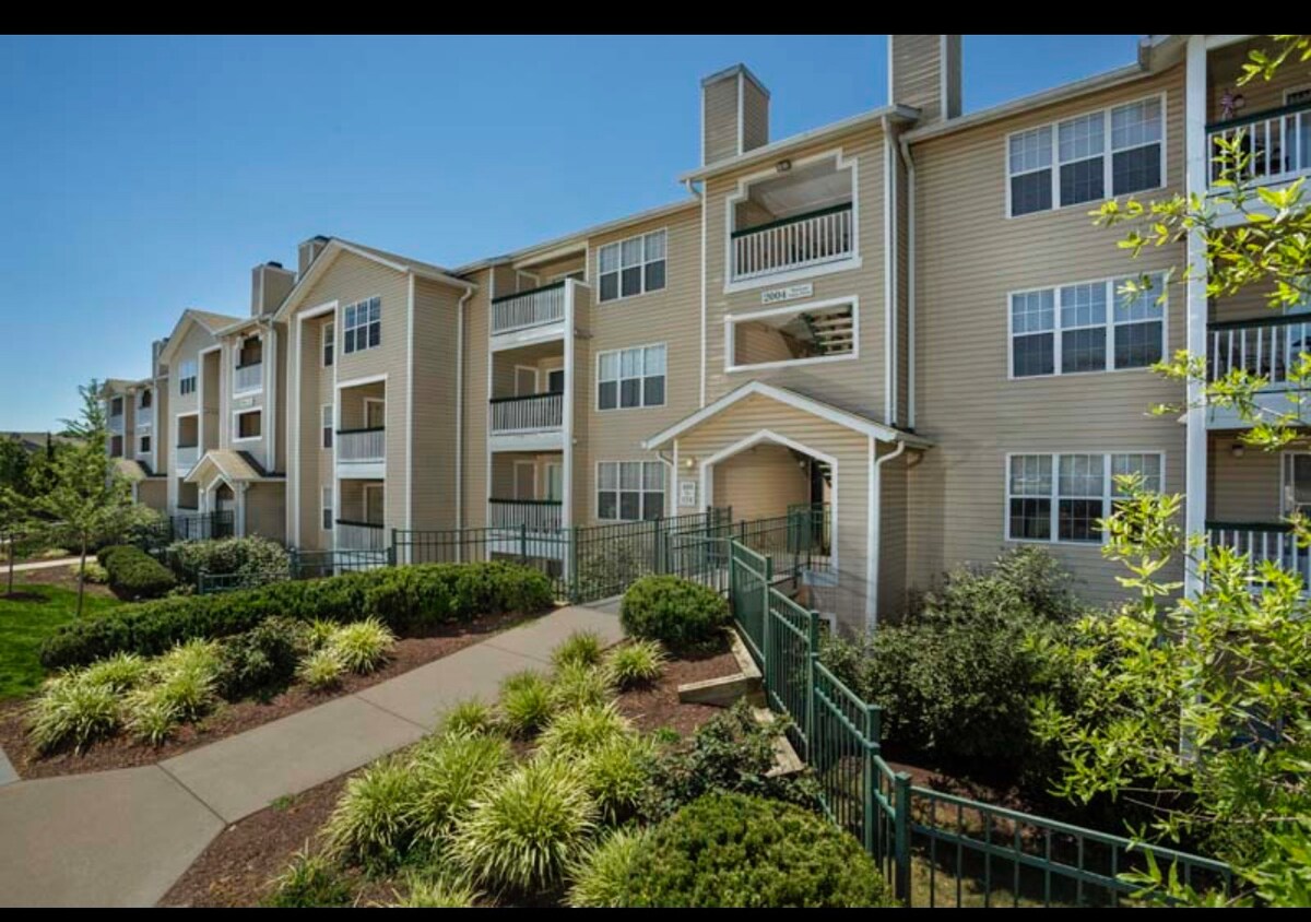 A well-maintained apartment complex is showcased with several balconies along the beige exterior. Lush landscaping, including shrubs and ornamental grasses, frames the walkway leading to the entrance, while clear blue skies provide a bright backdrop.