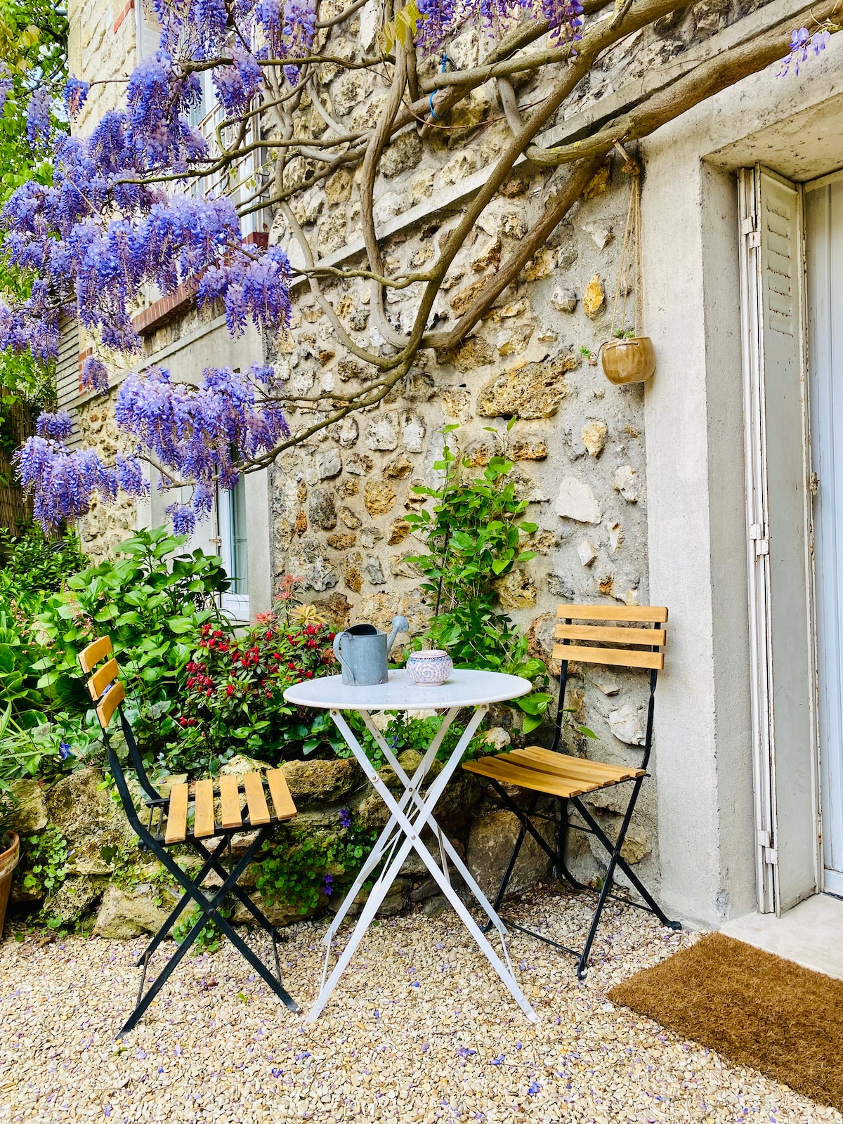 An outdoor seating area is displayed, featuring a white round table surrounded by two wooden chairs. Lush greenery and blooming wisteria create a colorful backdrop against the stone wall. The space invites guests to enjoy the garden ambiance.