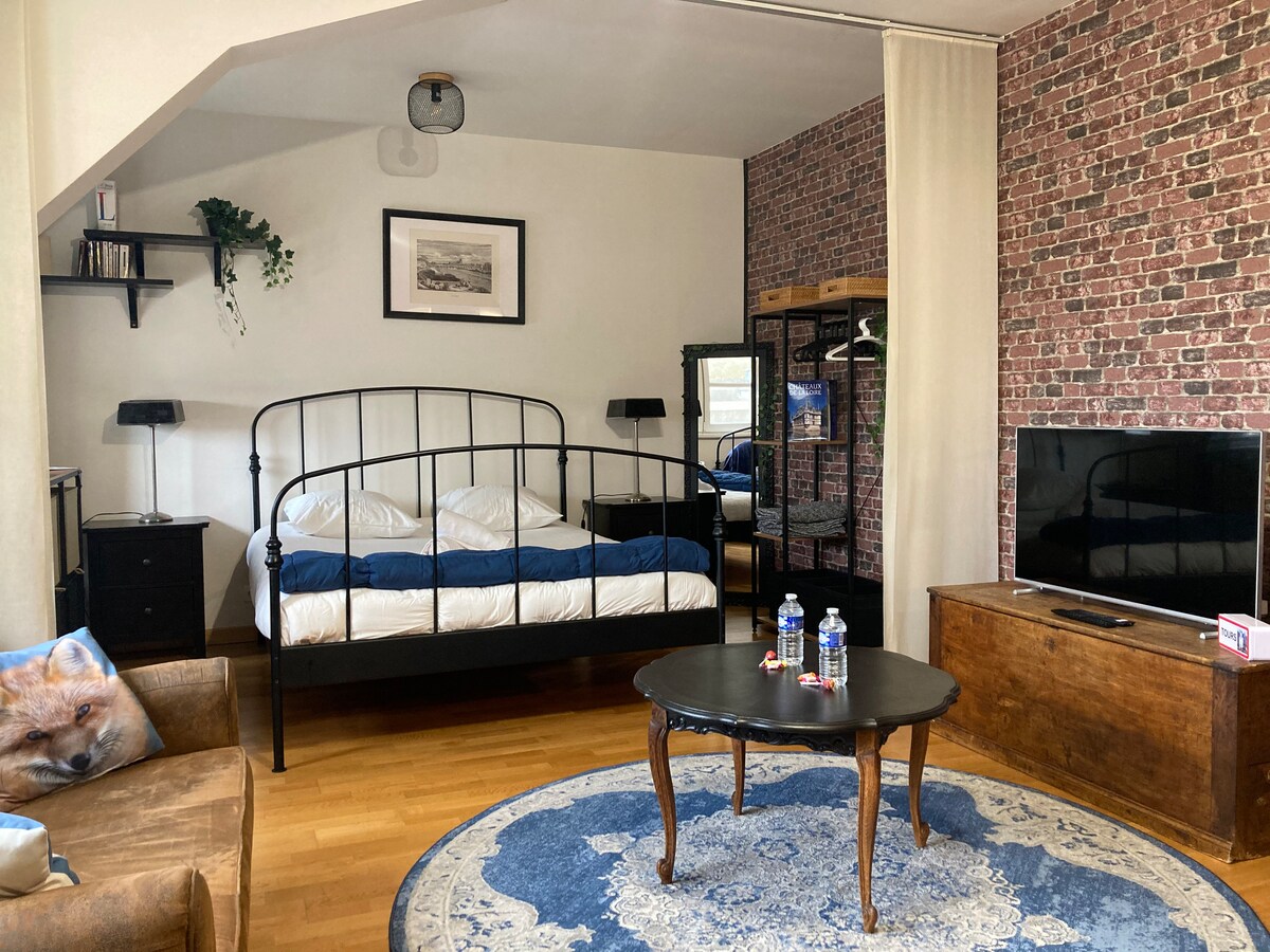 The space features a cozy living area adjacent to a sleeping area. A stylish bed with a minimalist design is set against a brick-patterned wall. A dark table is positioned at center, accompanied by two bottles of water. Warm wood flooring complements the inviting atmosphere.