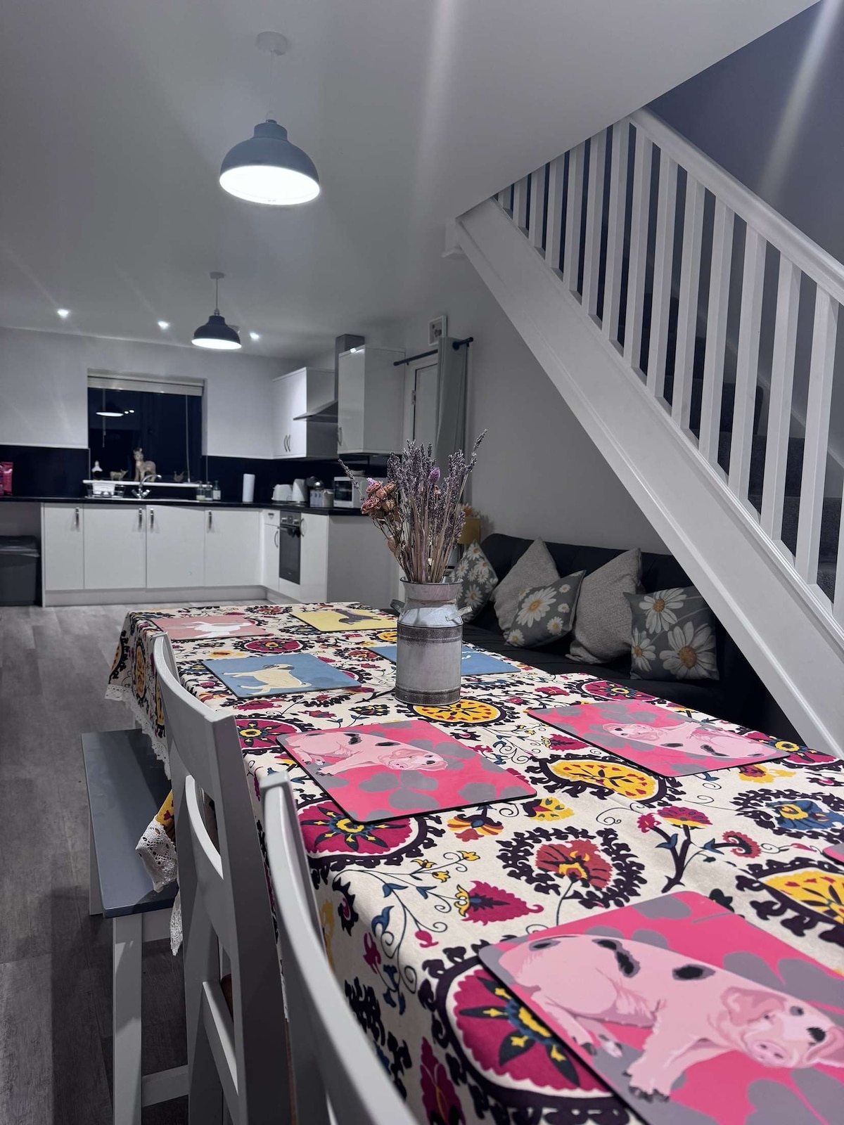 A dining area features a long table set with colorful placemats, surrounded by light grey chairs. A collection of lavender is displayed in a vase at the center. The space is illuminated by pendant lights, and the kitchen is visible in the background.