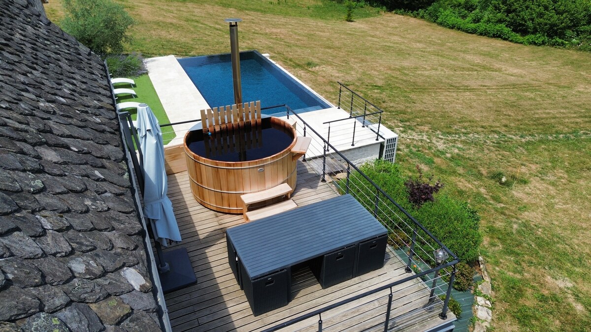 An aerial view captures a wooden nordic bath positioned on a spacious terrace beside a heated infinity pool. Sun loungers are arranged on the side, while green fields stretch out in the background, contributing to the serene outdoor setting.