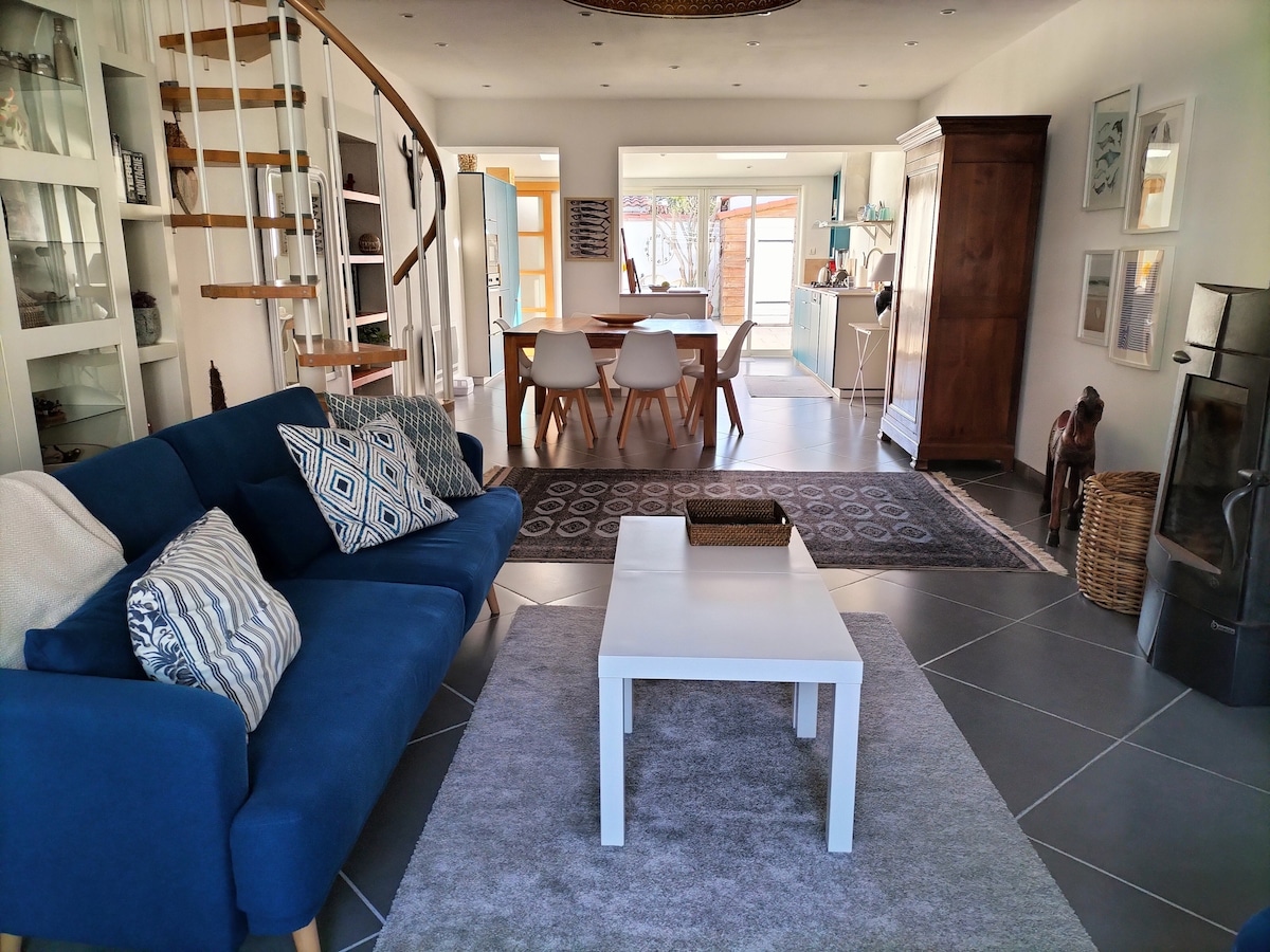 The living area features a comfortable blue sofa adorned with patterned cushions, facing a white coffee table. A round dining table is visible, surrounded by sleek white chairs. Natural light enters through large windows, illuminating the modern decor, including a staircase that leads to the upper level.