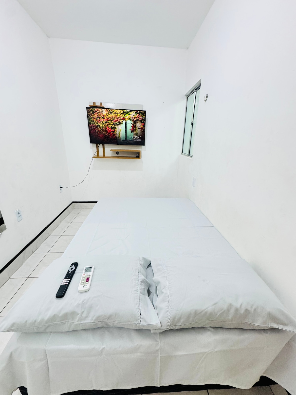 A simple sleeping area features a spacious bed covered with white bedding. A wall-mounted TV is visible above a small shelf, with remote control placed on the bed. Natural light enters through a window, illuminating the white walls and tiled floor.