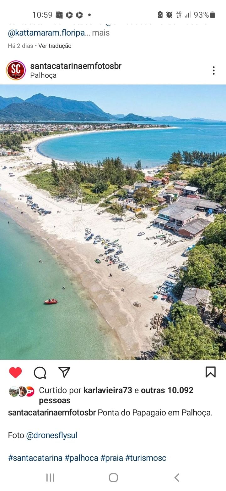 An aerial view captures a sweeping coastline with sandy beaches and gentle waves. Several vehicles and umbrellas are positioned along the shore, while lush greenery and mountains frame the landscape in the background, creating a serene seaside atmosphere.