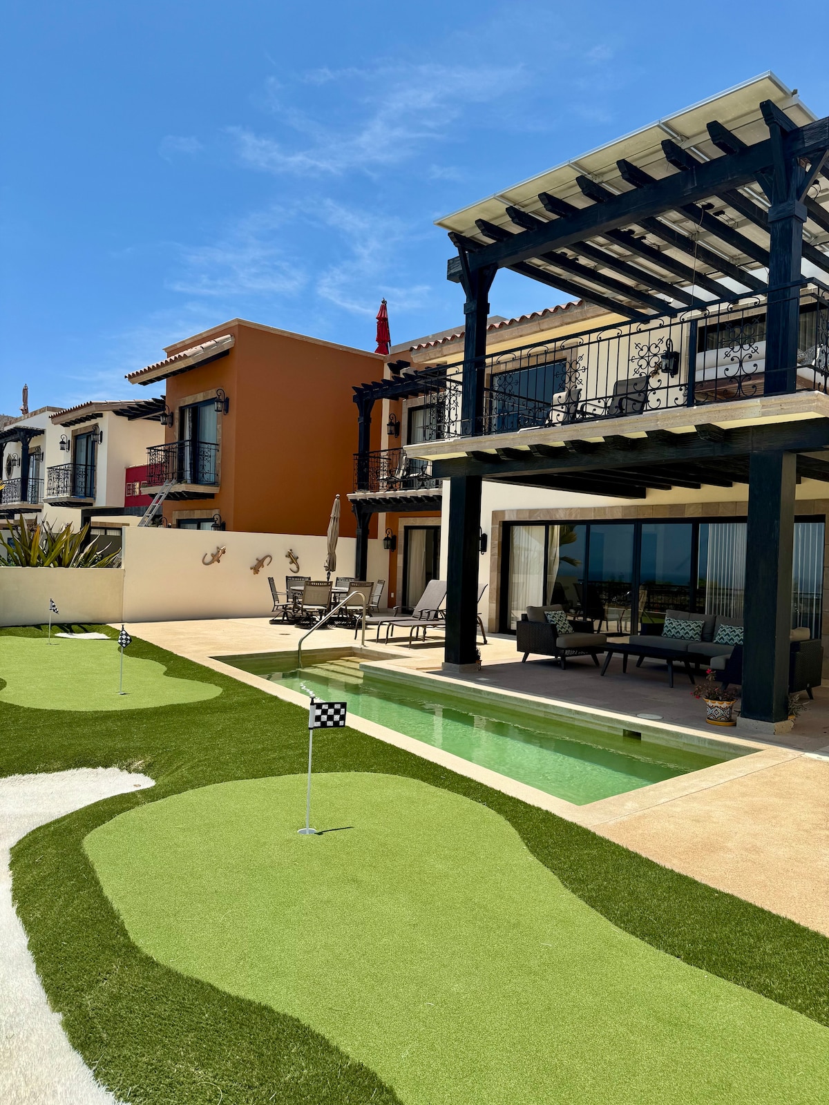 An outdoor area features a small private pool adjacent to a putting green with flags. Comfortable seating is provided under a shaded patio with a view of the two-story building in the background, showcasing a blend of architectural styles.