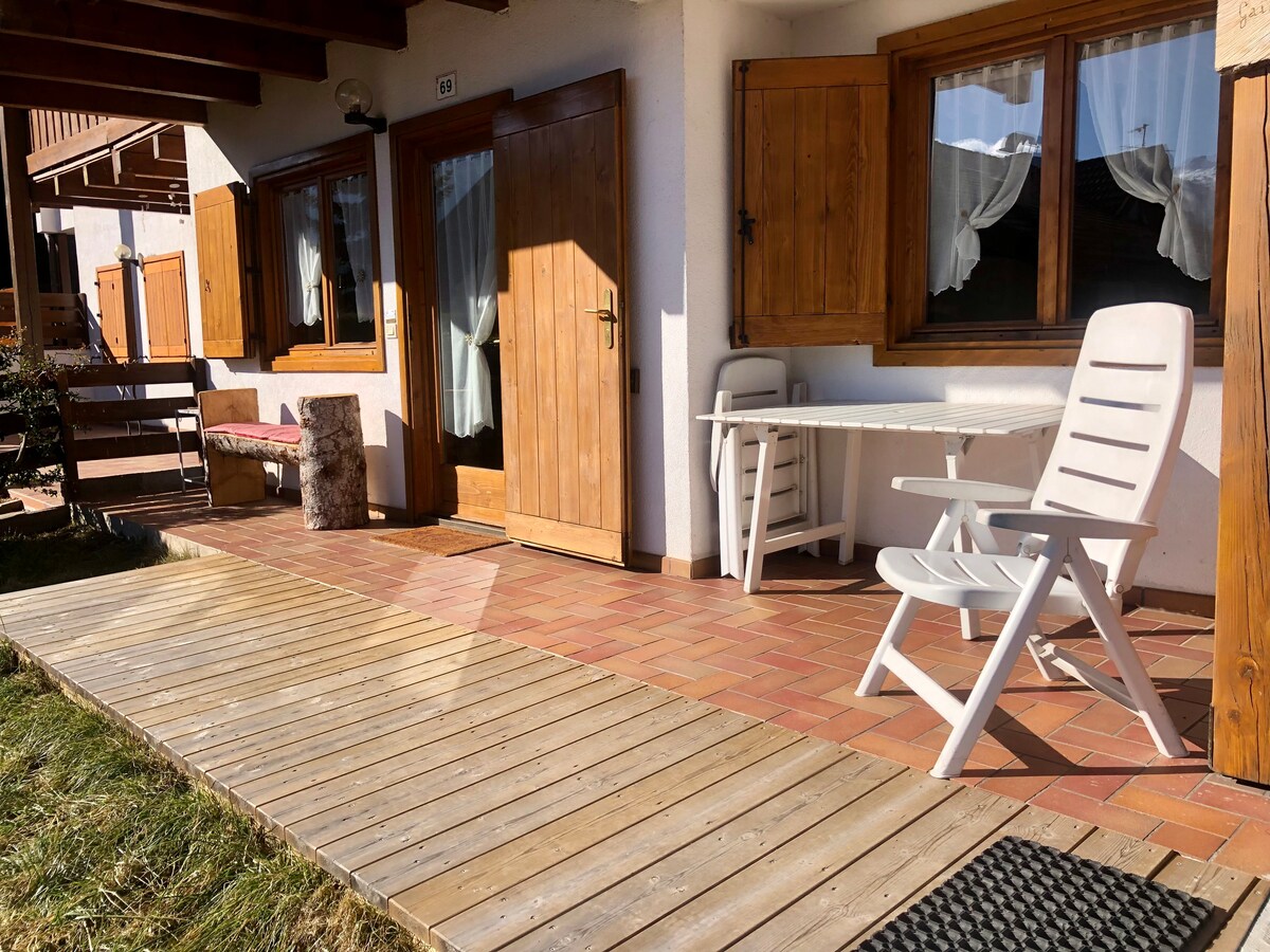 An outdoor patio features a wooden deck with a mix of brick flooring. White outdoor furniture, including a table and two chairs, is positioned near the entrance. Brown wooden shutters adorn the windows, and grass surrounds the patio area.