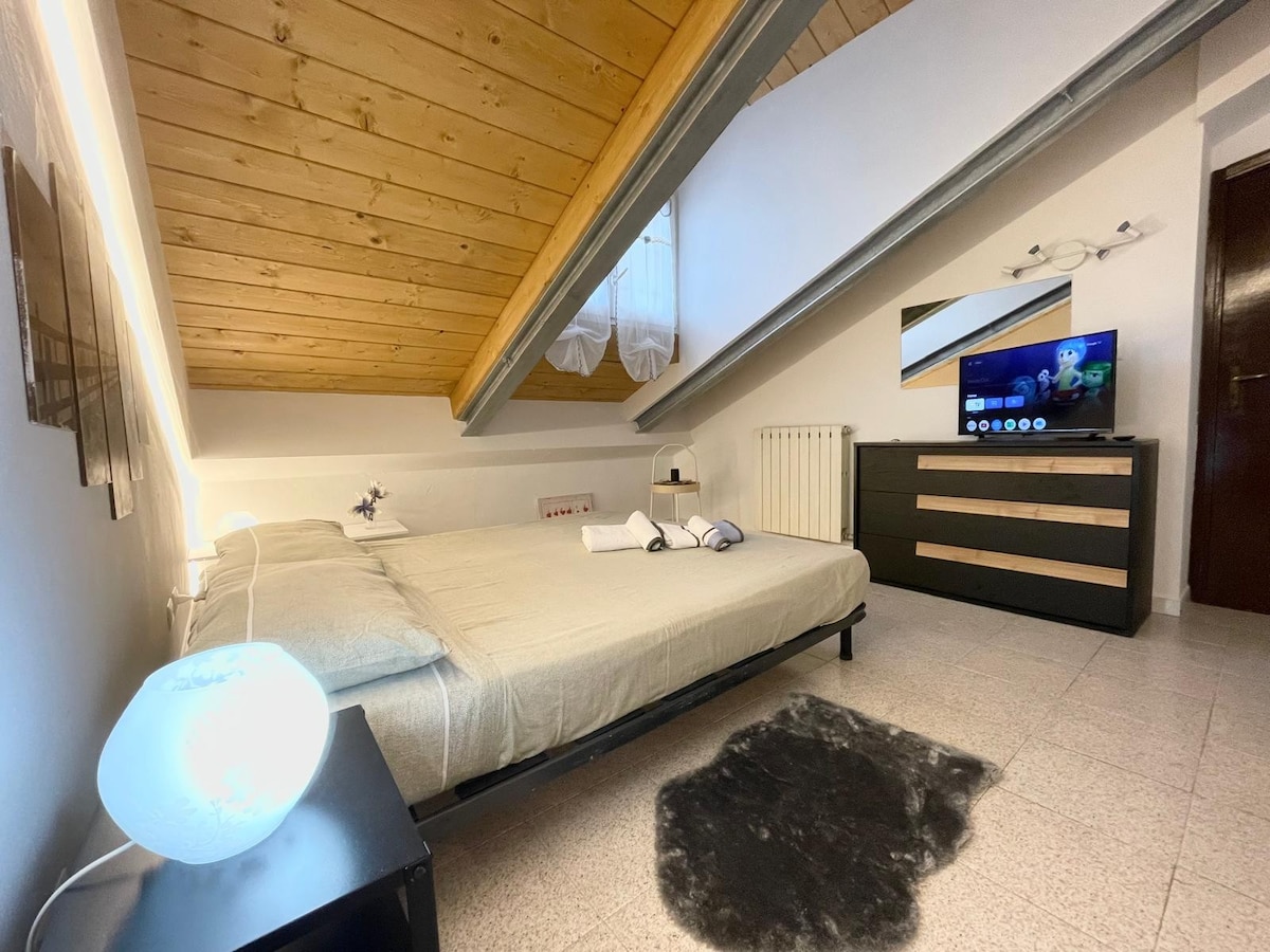 A serene bedroom features a spacious bed with crisp linens, positioned beneath a sloped wooden ceiling. A flat-screen television is mounted on the wall opposite the bed. Soft lighting highlights the room, while a small bedside table adds to the functional decor.
