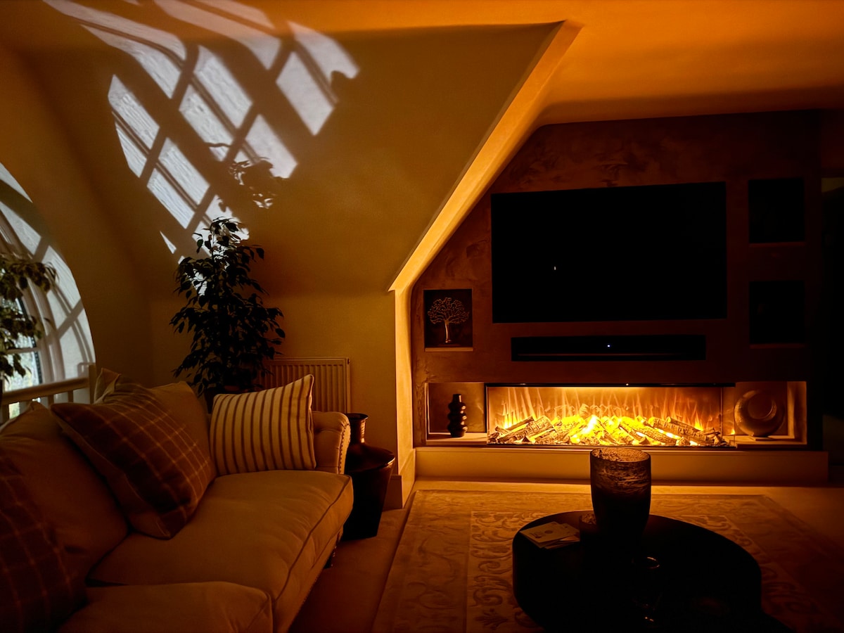 A cozy living room is highlighted by soft ambient lighting, featuring a spacious sofa adorned with patterned cushions. A modern fireplace is positioned below a large flat-screen television, while a decorative plant adds a touch of nature to the space. Light patterns from the windows create a calming atmosphere.