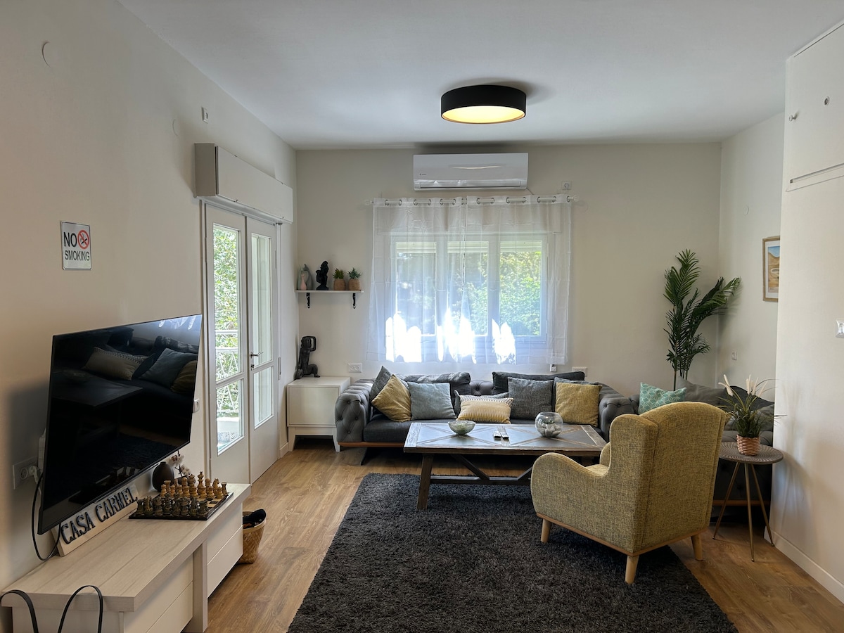 A spacious living area features a large, comfortable seating arrangement with a coffee table at its center. Natural light filters through large windows, illuminating the room. A flat-screen TV is positioned across from the seating, and a decorative plant adds a touch of greenery.