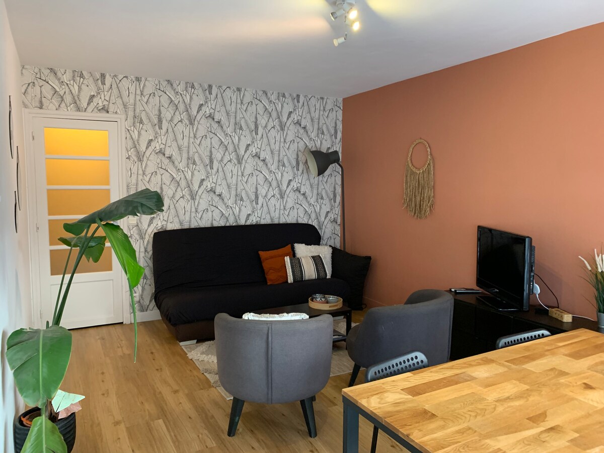 A spacious living area features a dark sofa, complemented by patterned cushions, and two armchairs. A medium-sized wooden table sits in front of a low black cabinet holding a television. Greenery from a nearby plant adds a touch of freshness to the modern decor.