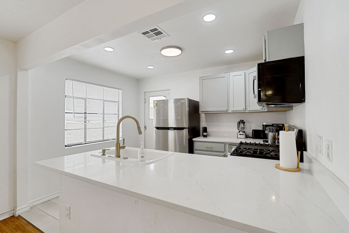 A modern kitchen features a spacious marble countertop with a sink and contemporary faucet. Stainless steel appliances, including a refrigerator, oven, and microwave, are visible. Ample cabinet space is provided, and natural light enters through a window, illuminating the clean design of the room.