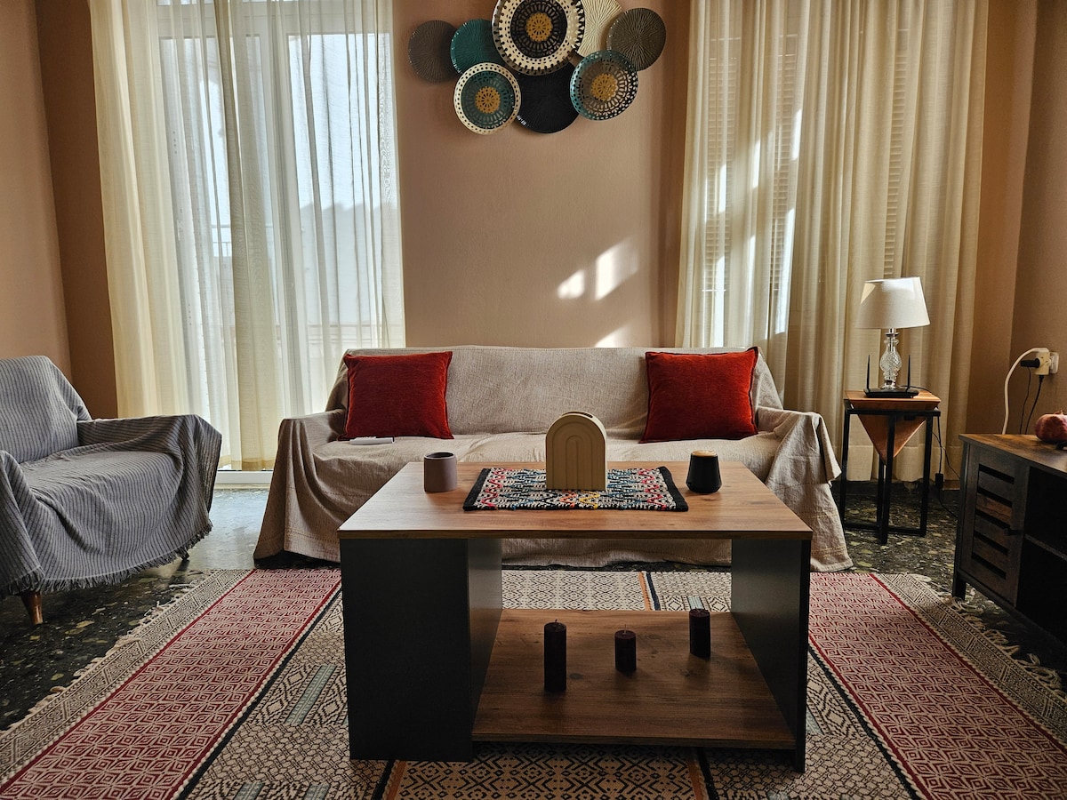 A cozy living space features a light-colored sofa with red accent pillows and a patterned rug beneath a wooden coffee table. Natural light filters through sheer curtains, illuminating decorative wall art and a small lamp on an adjacent side table.