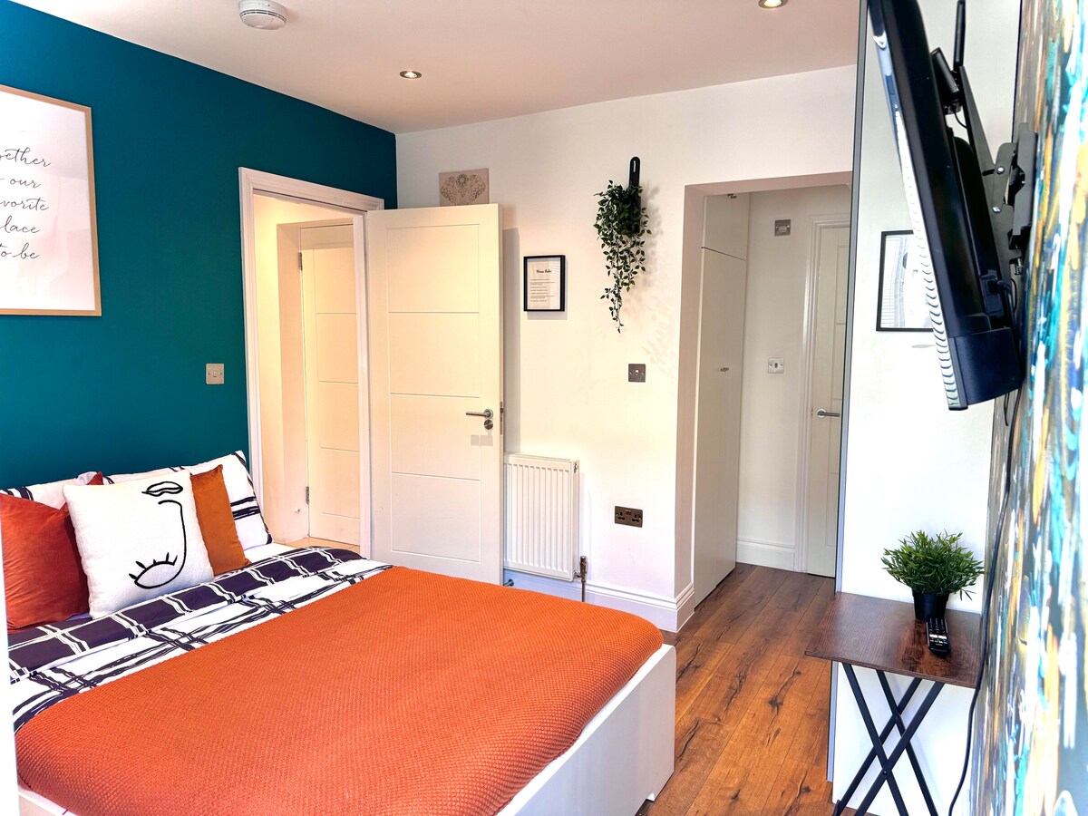 A studio apartment is presented with a double bed featuring a vibrant orange bedspread and decorative pillows. A television is mounted on the wall opposite a small table. The room's blue accent wall adds a modern touch, while light streaming in from the doorway illuminates the space.