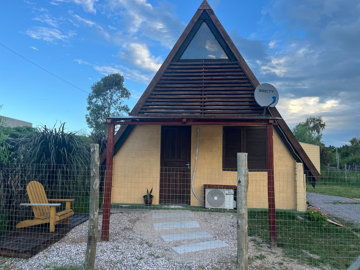 An A-frame cabin features a triangular roof and large front window, framed by lush greenery. A small pathway leads to the entrance, which is flanked by a porch and a seating area. Satellite dish is mounted for connectivity.