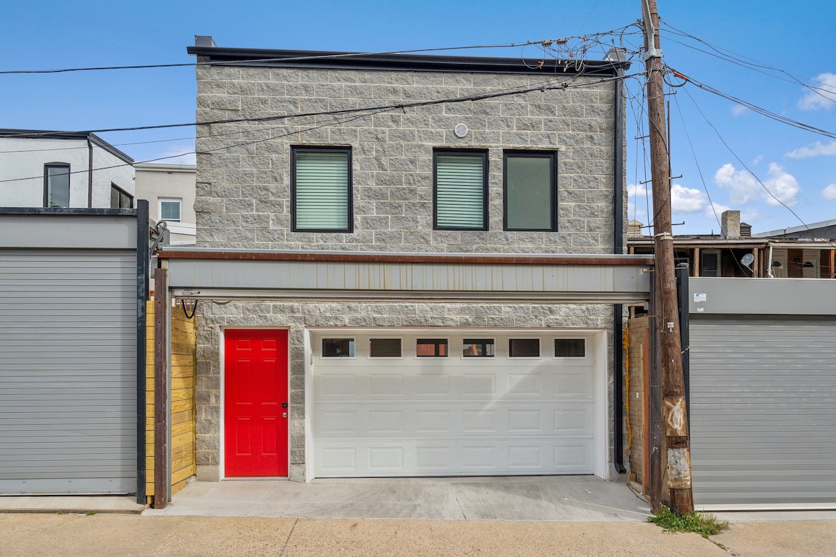 The exterior of the new construction apartment features a modern design with a striking red door and large garage door. Gray stone walls complement the clean lines of the building. Two small windows allow natural light into the upper level, set against a clear blue sky.