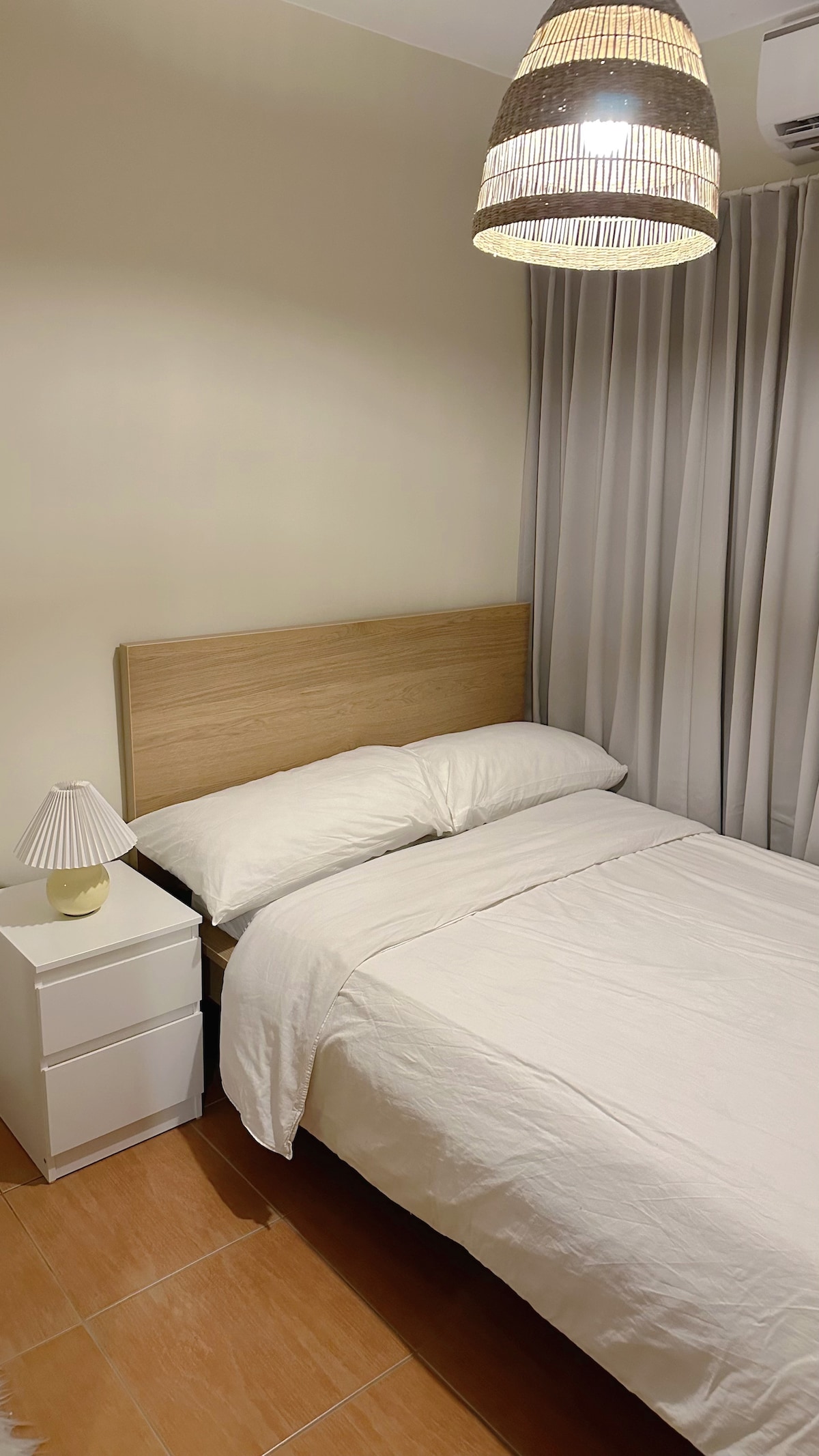 A well-lit bedroom features a cozy bed with neutral-colored linens, complemented by a wooden headboard. A white bedside table holds a small lamp, while soft curtains frame the window, allowing natural light to enter the space.