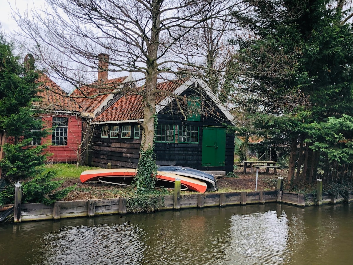 A charming exterior of a vintage house is surrounded by lush greenery and trees. Canoes are visible alongside the water's edge, hinting at recreational opportunities. The building's unique architectural elements showcase a combination of red brick and dark wood, while a green door adds a vibrant touch.