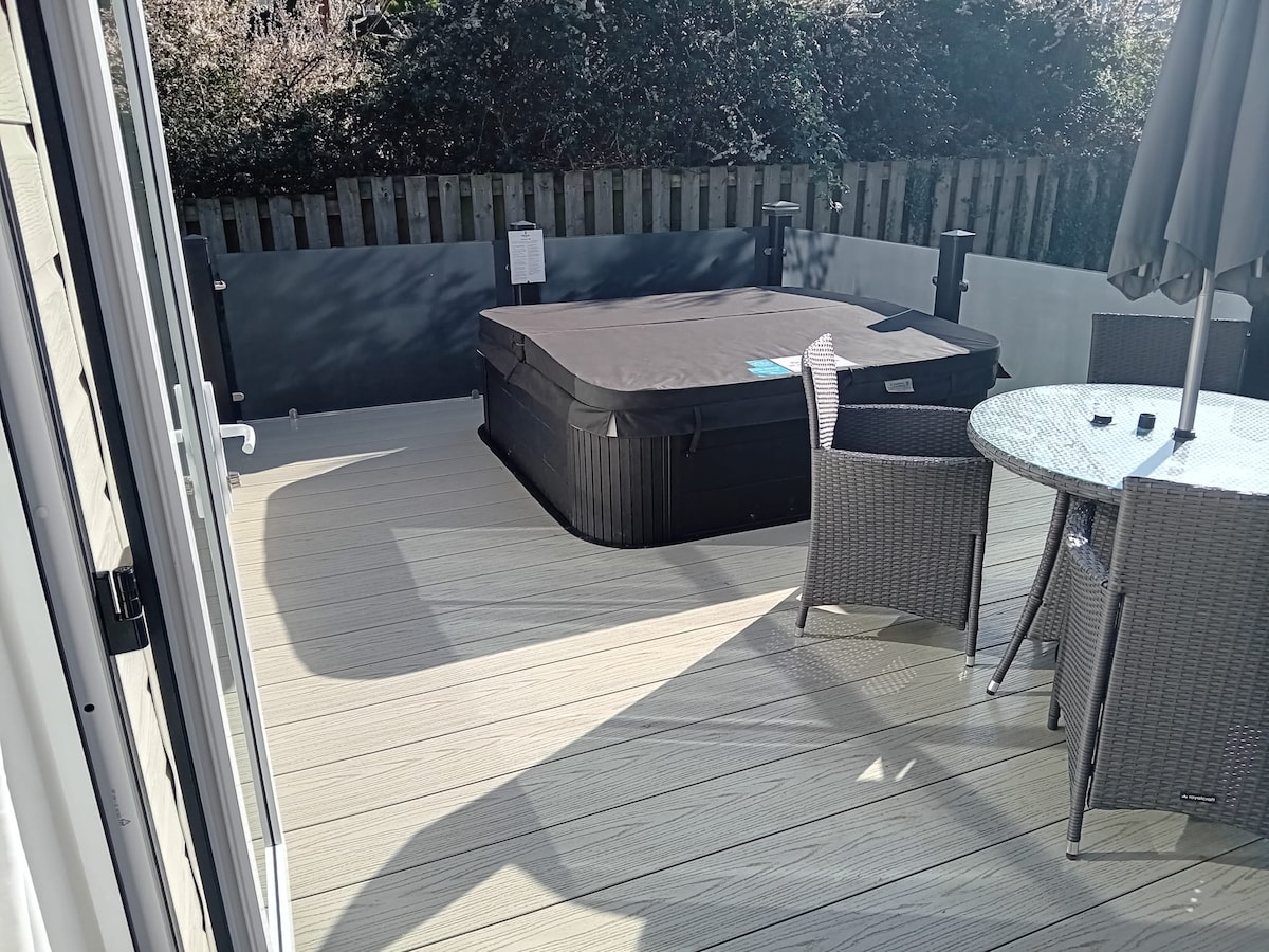A spacious decking area features a hot tub with a covered top and a round dining table surrounded by four woven chairs. Natural light casts soft shadows across the decking, creating a bright and inviting outdoor space.