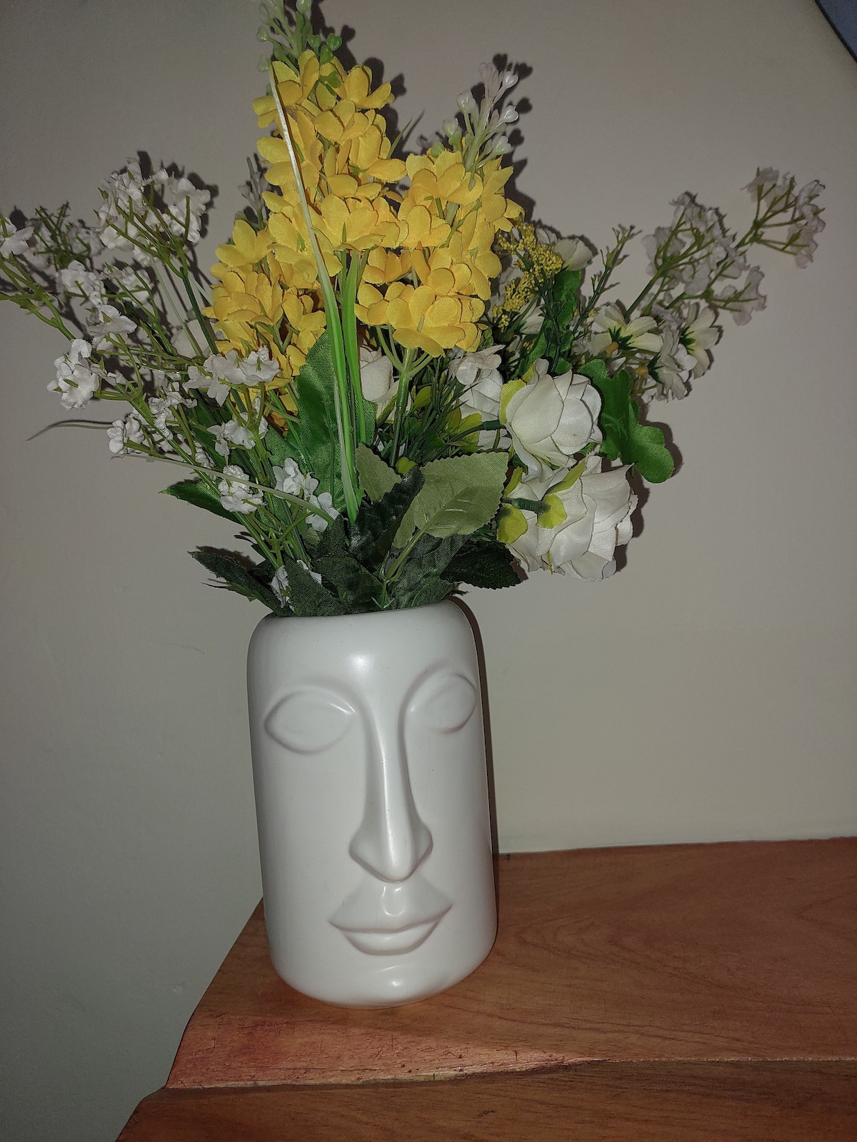 A decorative vase with a face design holds a vibrant arrangement of yellow flowers alongside white blossoms. The vase is positioned on a wooden surface, adding a touch of nature to the interior.