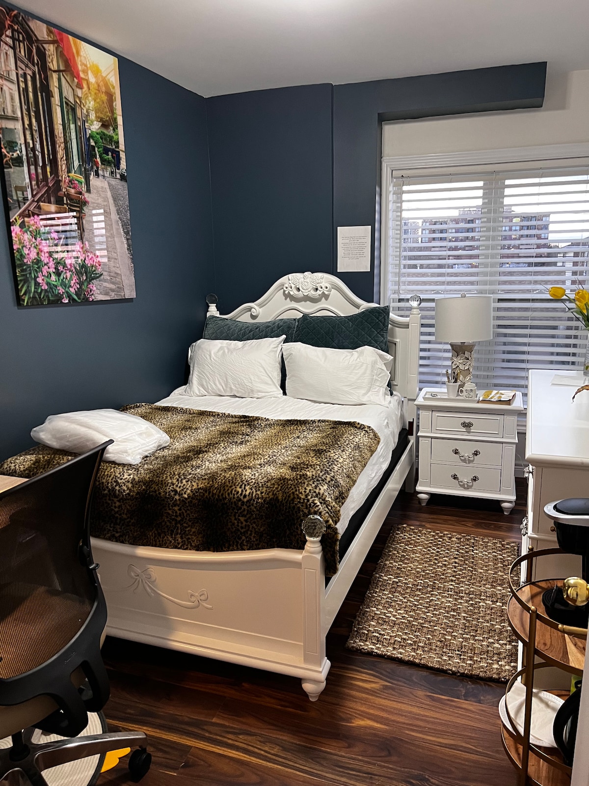 A cozy bedroom features a white bed with a patterned blanket and crisp linens. A bedside lamp is placed next to a white dresser. A large window is fitted with blinds, allowing natural light to brighten the room. A framed artwork adorns the wall, adding charm.