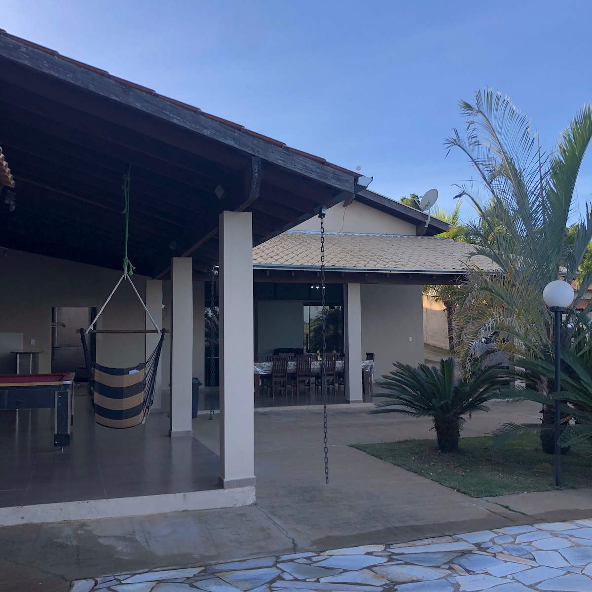 A spacious outdoor area is presented, featuring a covered terrace adjoining a modern dining space. A hanging swing chair is visible, along with a well-maintained garden of palms and other greenery. The floor is paved, complementing the structure's contemporary design.