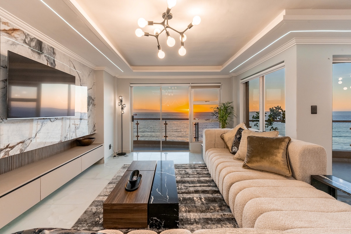 A spacious living area features a large, curved sectional sofa complemented by decorative cushions. A contemporary coffee table is central to the room. Floor-to-ceiling windows frame a stunning sunset over the ocean, while a modern light fixture adds an elegant touch to the ceiling.