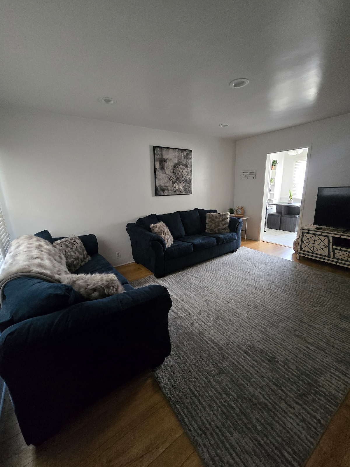 A bright living area is furnished with two dark blue sofas adorned with decorative pillows. A large area rug covers the wooden floor, and natural light enters through a doorway leading to another room, creating a welcoming space for relaxation.