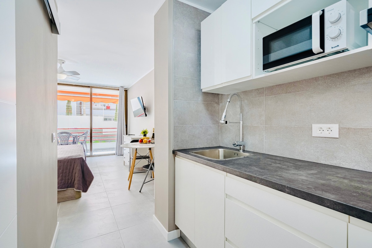 A modern kitchen area is combined with a living space, featuring a sleek countertop and stainless-steel sink. A microwave and additional kitchen appliances are visible. Light filters through a glass door, leading to an outdoor space with a dining table and chairs.