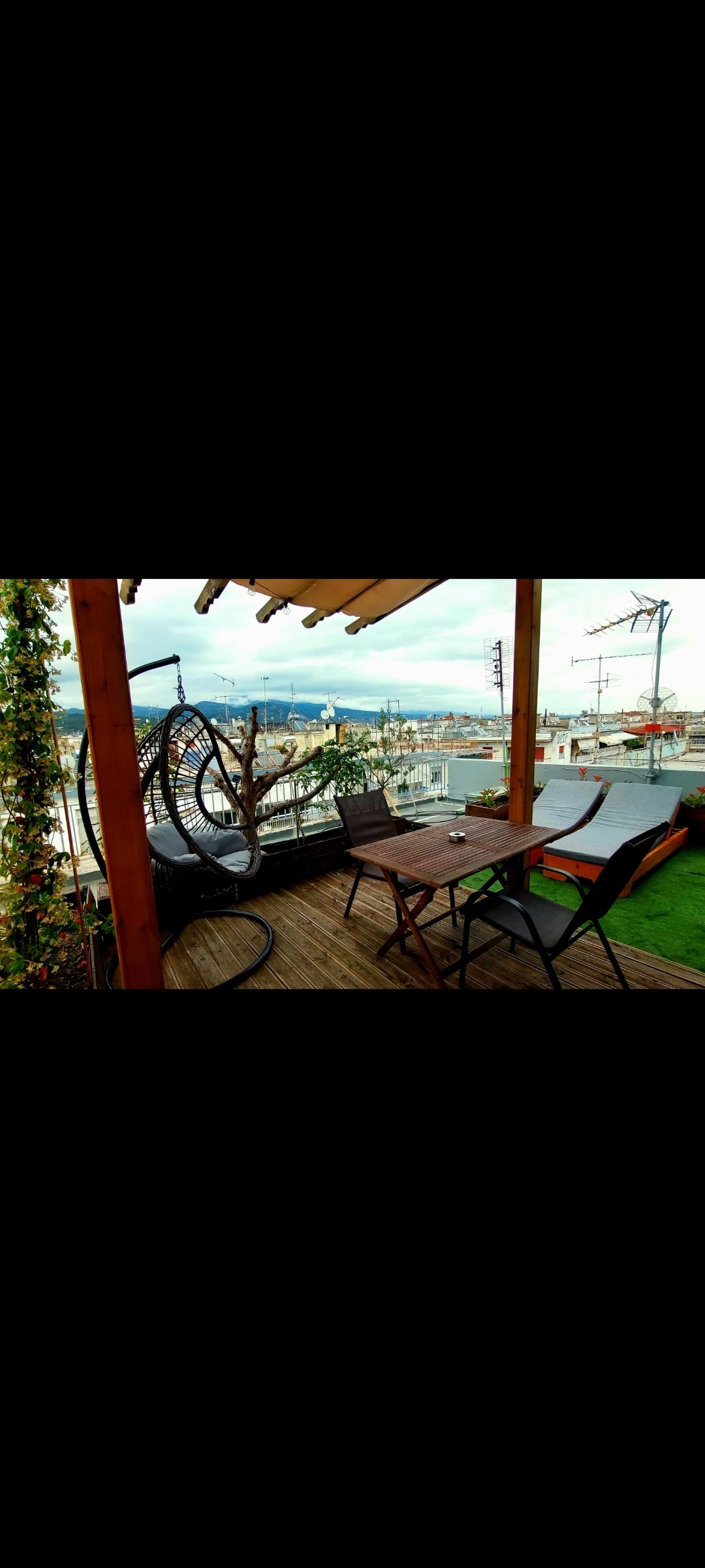 A wooden deck is showcased, featuring a hanging chair and a dining table with four seats. A view of the city skyline is visible in the background, with greenery and outdoor decor enhancing the space.