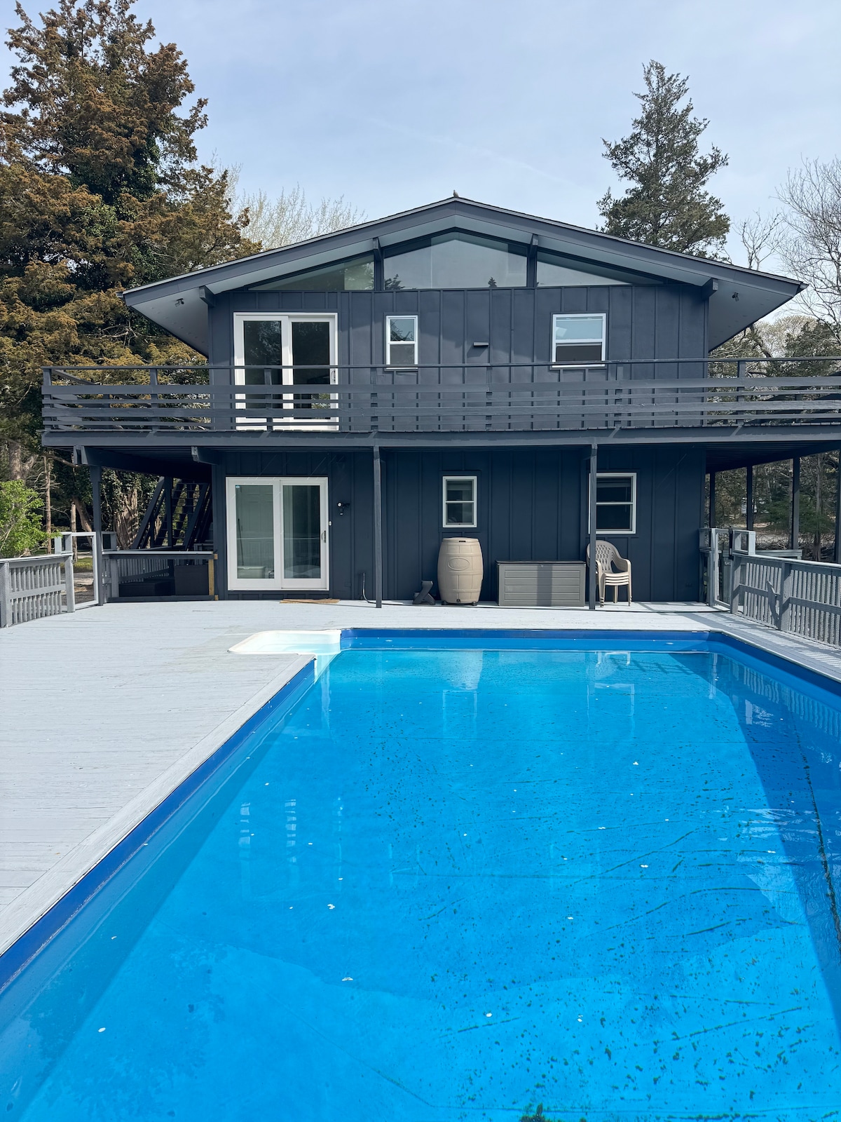 The two-story home is framed by a spacious deck and a blue swimming pool. Large windows provide a view into the house, while a light-colored seating area is visible on the deck. The exterior is painted in a dark hue, creating a modern aesthetic.