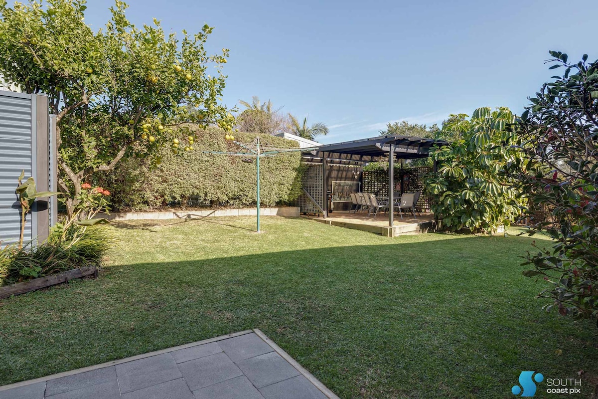 A spacious backyard is shown, featuring well-maintained grass and tropical foliage. A covered area with seating is visible, along with a clothesline and garden path. Landscaping elements create a serene outdoor environment suitable for relaxation or family activities.