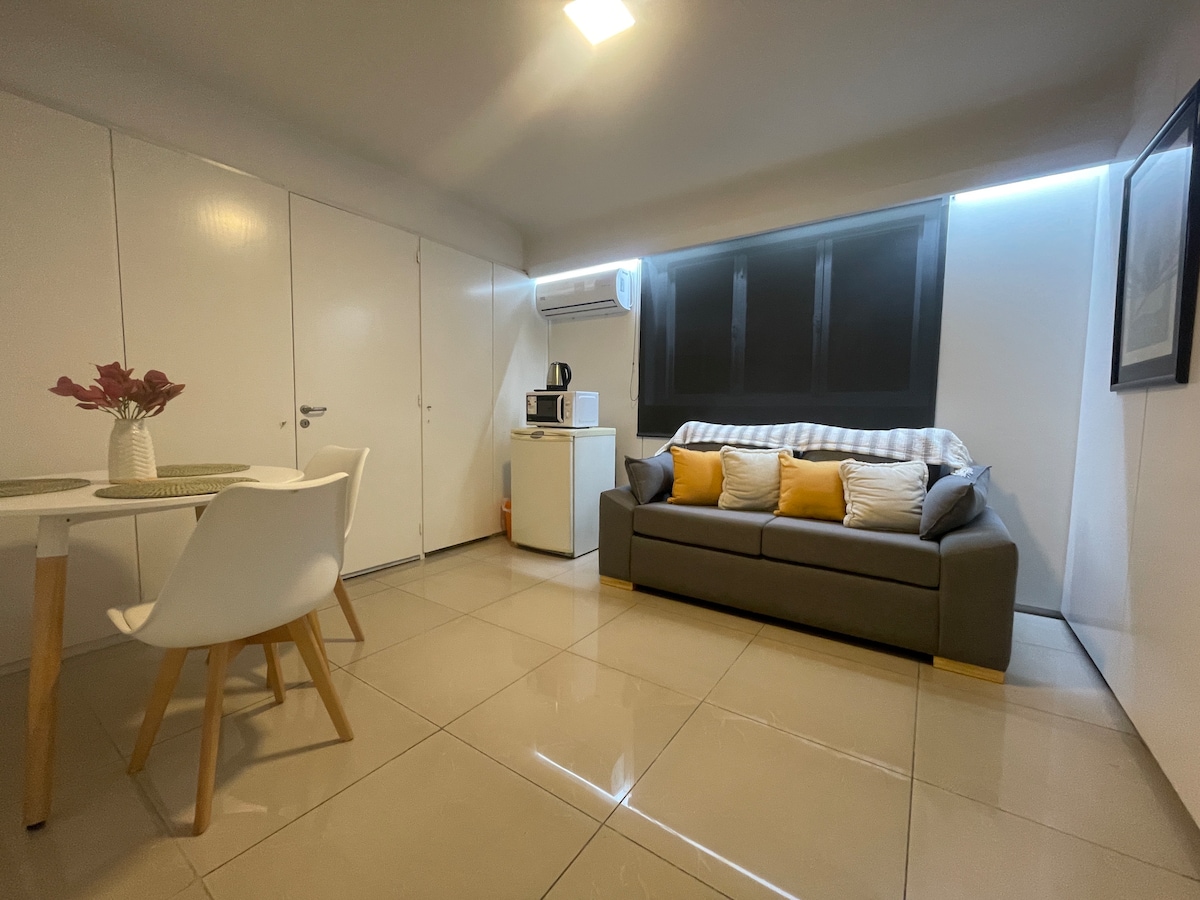 A cozy living area is present, featuring a grey sofa adorned with yellow cushions. A dining table with white chairs sits nearby. The kitchenette includes a microwave and a small refrigerator. Natural light is filtered through a large window, complemented by soft, ambient lighting.