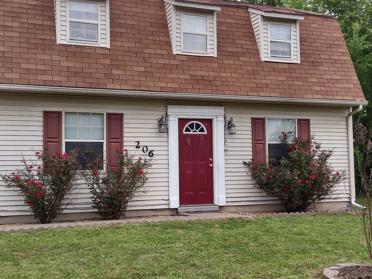 The exterior of the home features a welcoming design with a red front door and white shutters. Lush greenery surrounds the entrance, complemented by flowering bushes on either side. A neatly manicured lawn enhances the inviting appearance of the property.