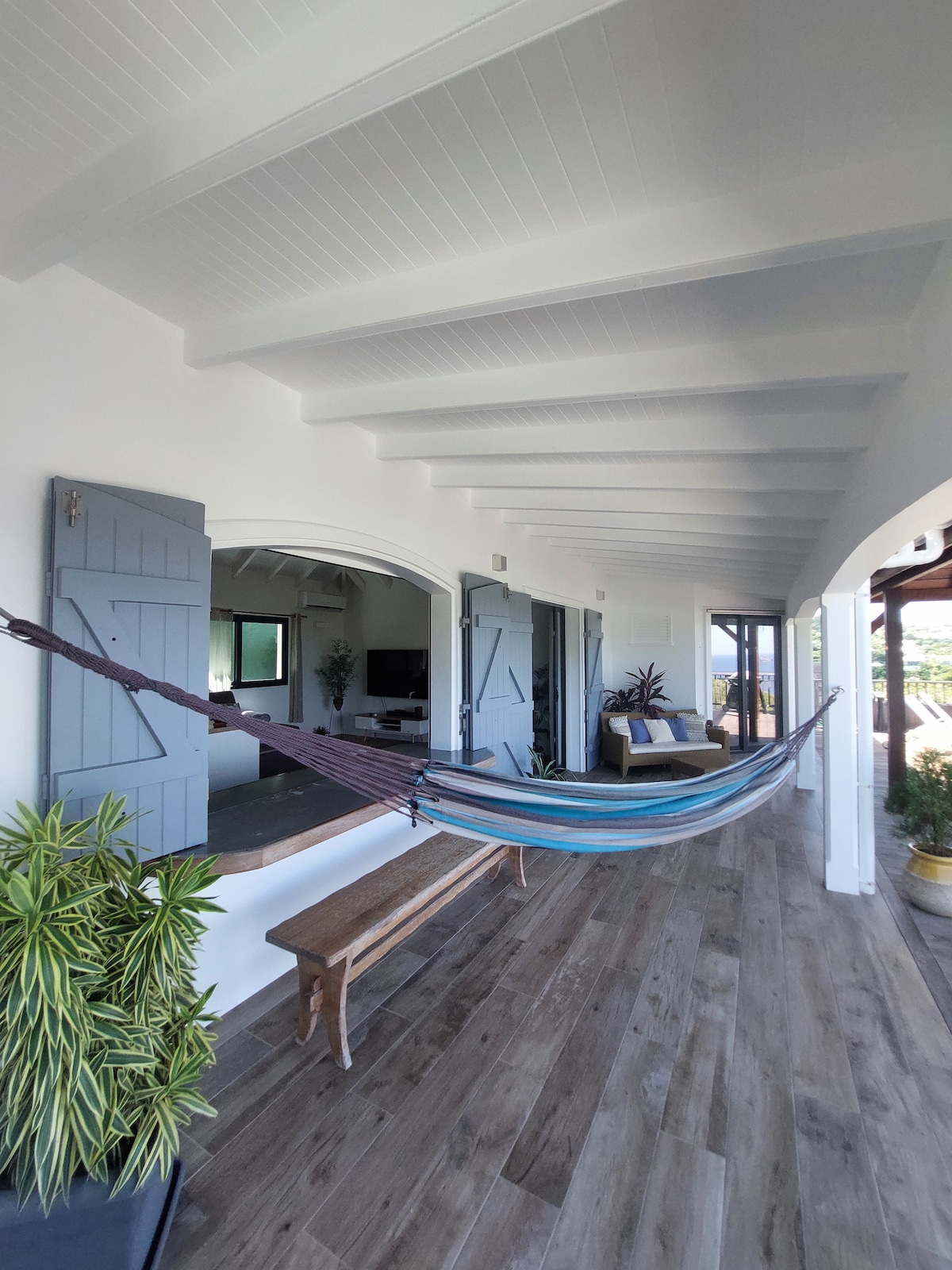 A covered terrace space includes a colorful hammock suspended between two walls. The airy area features wooden flooring and large arched openings leading to the living room. Green plants add a natural touch, enhancing the serene atmosphere with sunlight filtering in through the windows.