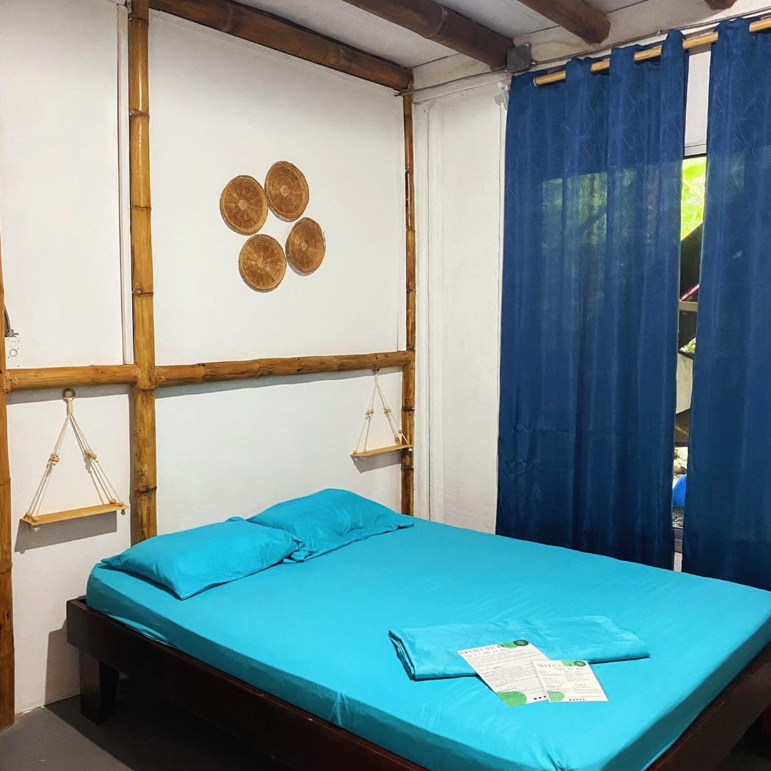 A private room features a spacious bed covered with a blue sheet. Two shelves are mounted on the bamboo walls, and a window with blue curtains offers natural light. Wall decorations add a touch of character, enhancing the tranquil setting.