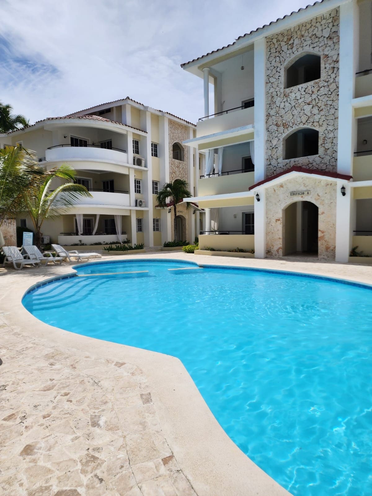 A clear blue swimming pool is surrounded by lounge chairs and tropical landscaping. The building features a stone facade and balconies, creating a harmonious outdoor space for relaxation. Sunlight reflects off the water, enhancing the inviting atmosphere of this leisure area.