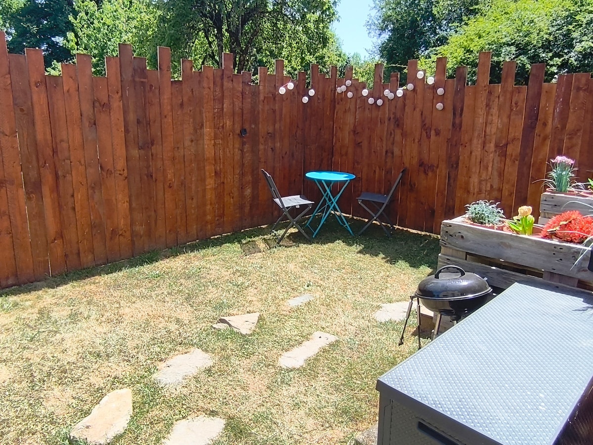 An enclosed outdoor area features a wooden fence and a patch of grass. A small blue table is accompanied by two folding chairs, while planters with flowers add color. A charcoal grill is present beside a storage unit, creating a cozy space for relaxation.
