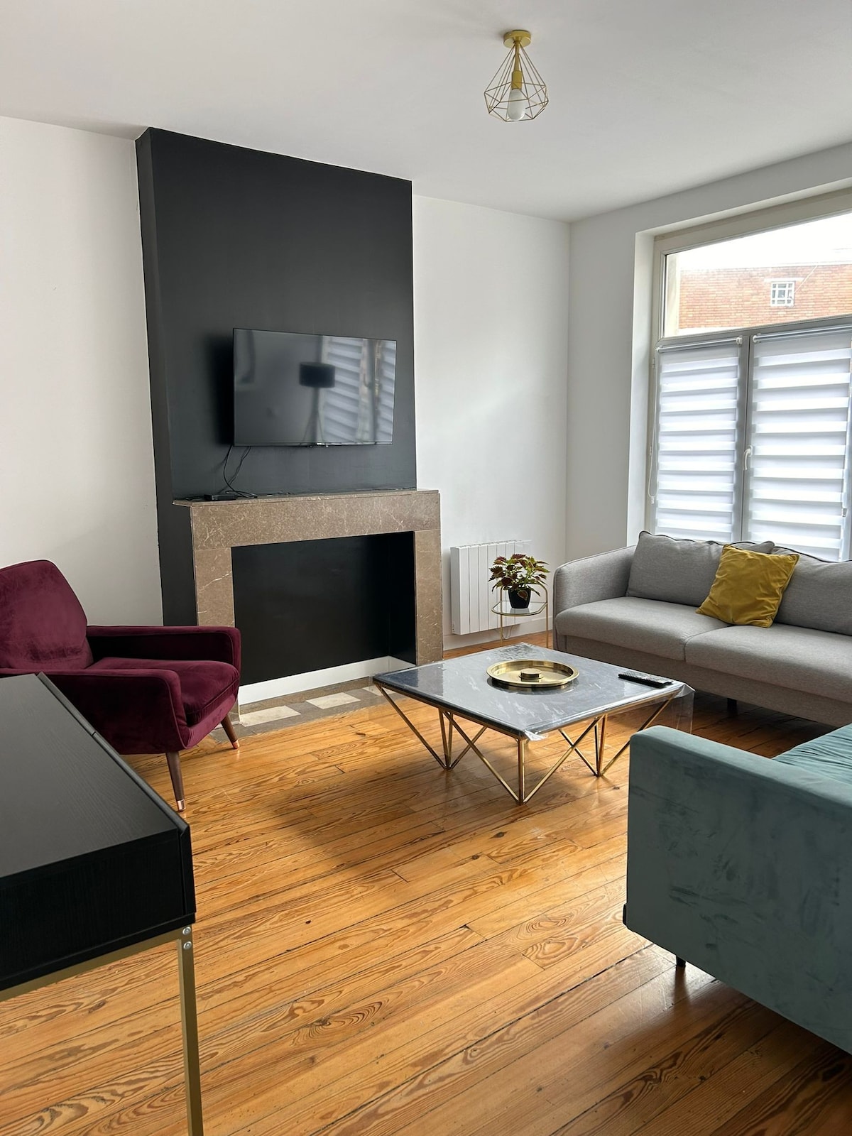 A bright living area features a sleek television mounted above a modern fireplace. A glass coffee table is positioned centrally, surrounded by a sofa and two armchairs in muted tones. Natural light enters through a large window with slatted shades, highlighting the warm wooden floor.