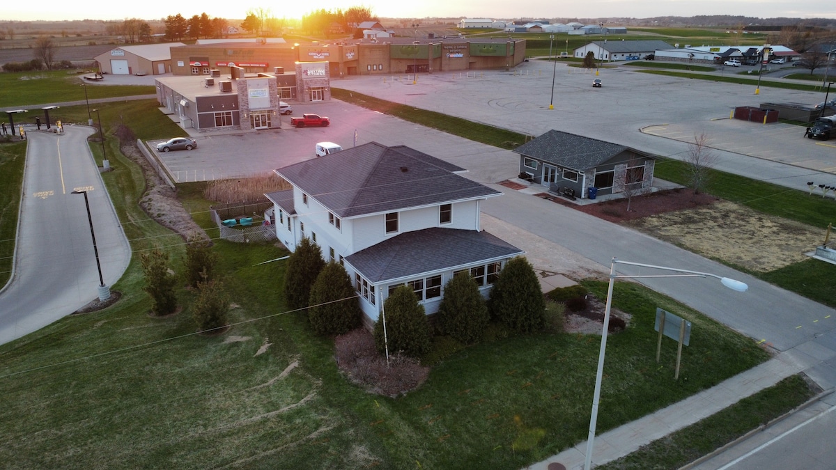 An aerial view captures a spacious two-story house surrounded by green grass and neatly trimmed shrubs. The house is positioned near commercial buildings and open parking areas, with a sunset casting warm light over the scene.